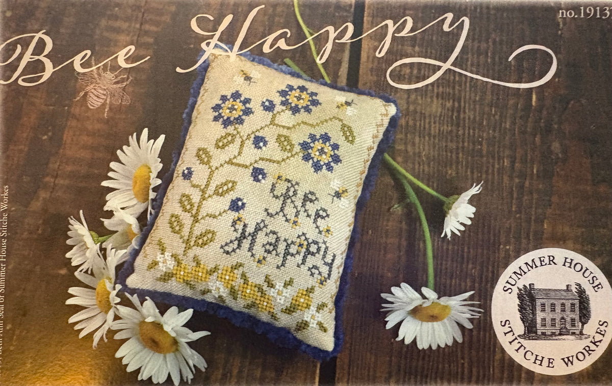 A square cross-stitch pillow with a blue and yellow floral design and the words 'Bee Happy' stitched in the center, displayed on a rustic wooden background with white daisies. The image includes 'Bee Happy' script at the top and a 'Summer House Stitches Works' logo at the bottom right.