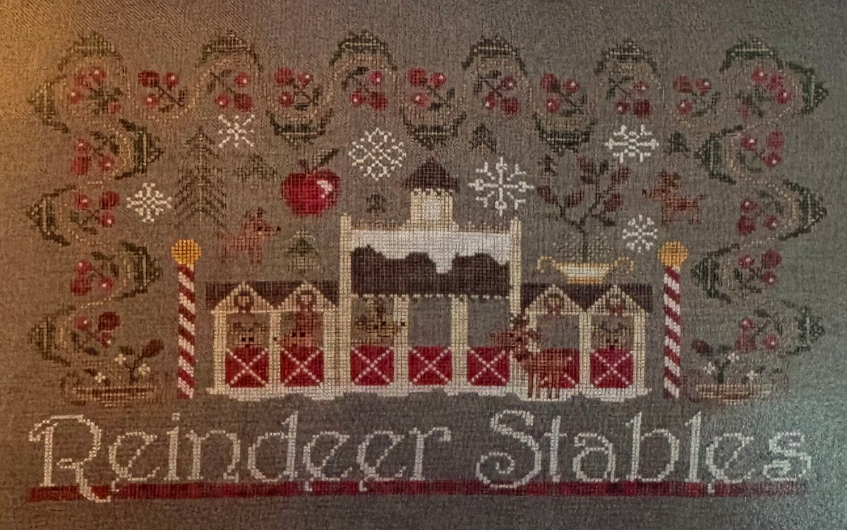 A needlepoint or cross-stitch style holiday design reading 'Reindeer Stables' on a gray fabric background, featuring a central stable with red barns, snowflakes, berries, trees, and reindeer motifs.