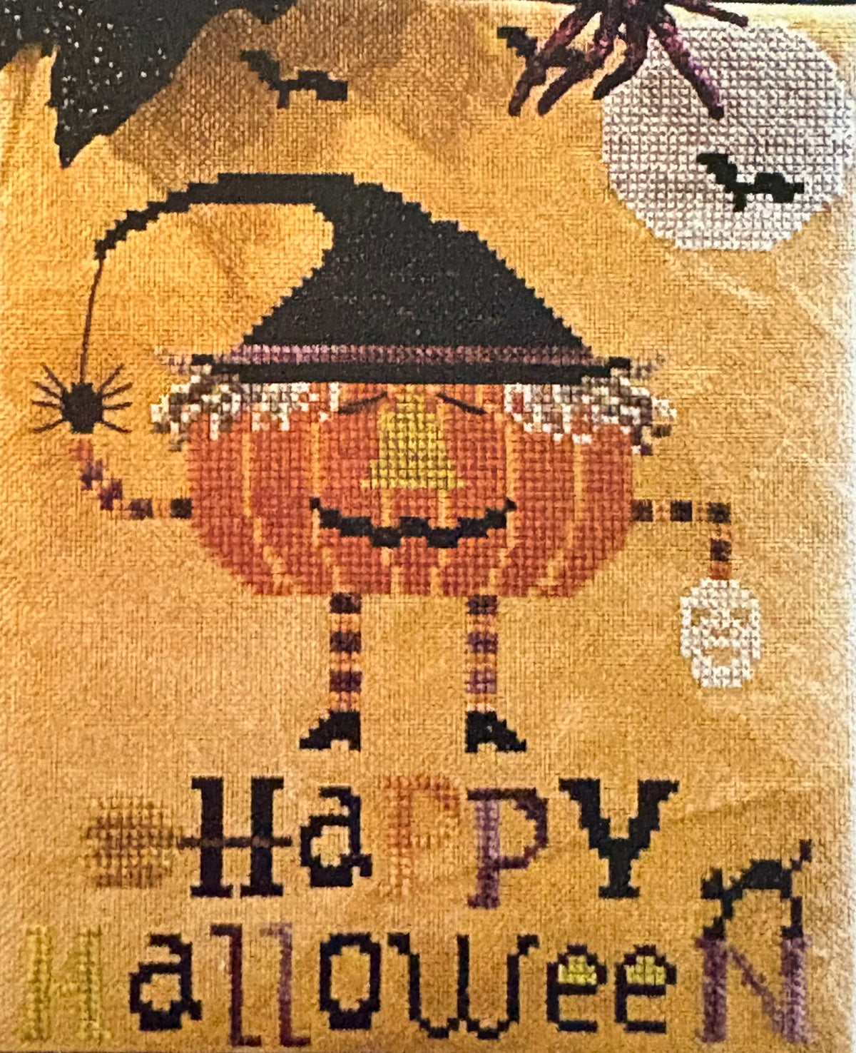 A close-up of a crocheted or knitted textile featuring a Halloween design: a smiling orange pumpkin with a black witch hat, black spider, bats, and the words “Happy Halloween” in multicolored lettering on a tan background.