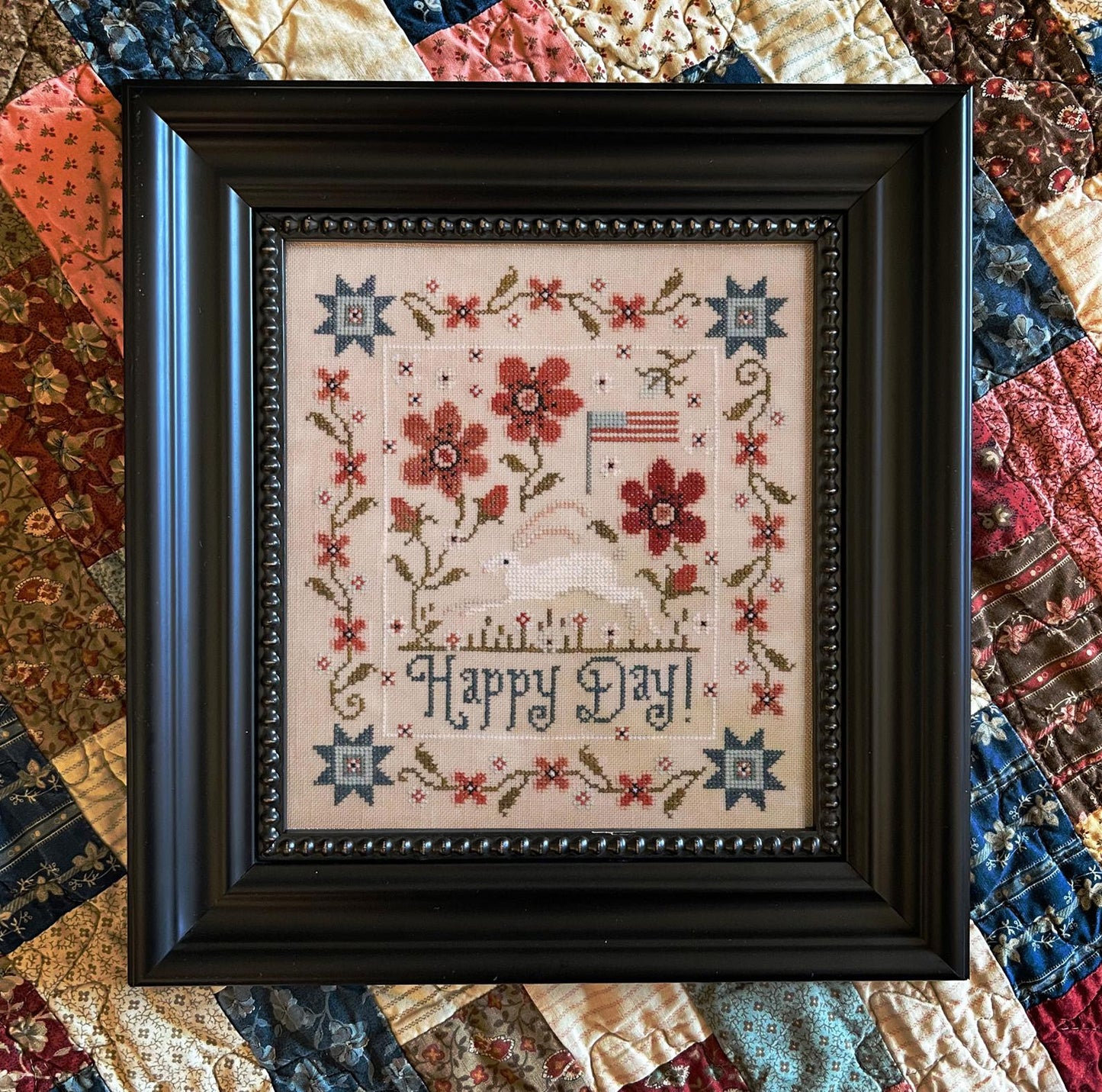 Framed cross-stitched piece with floral design and 'Happy Day!' text on a quilt background