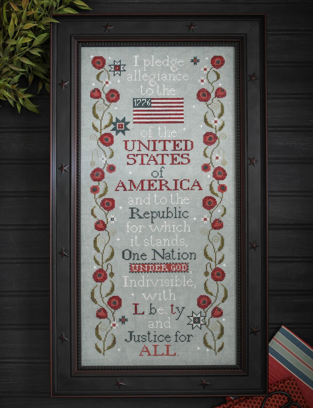 Framed cross-stitched piece with the Pledge of Allegiance and American flag on a wooden background.