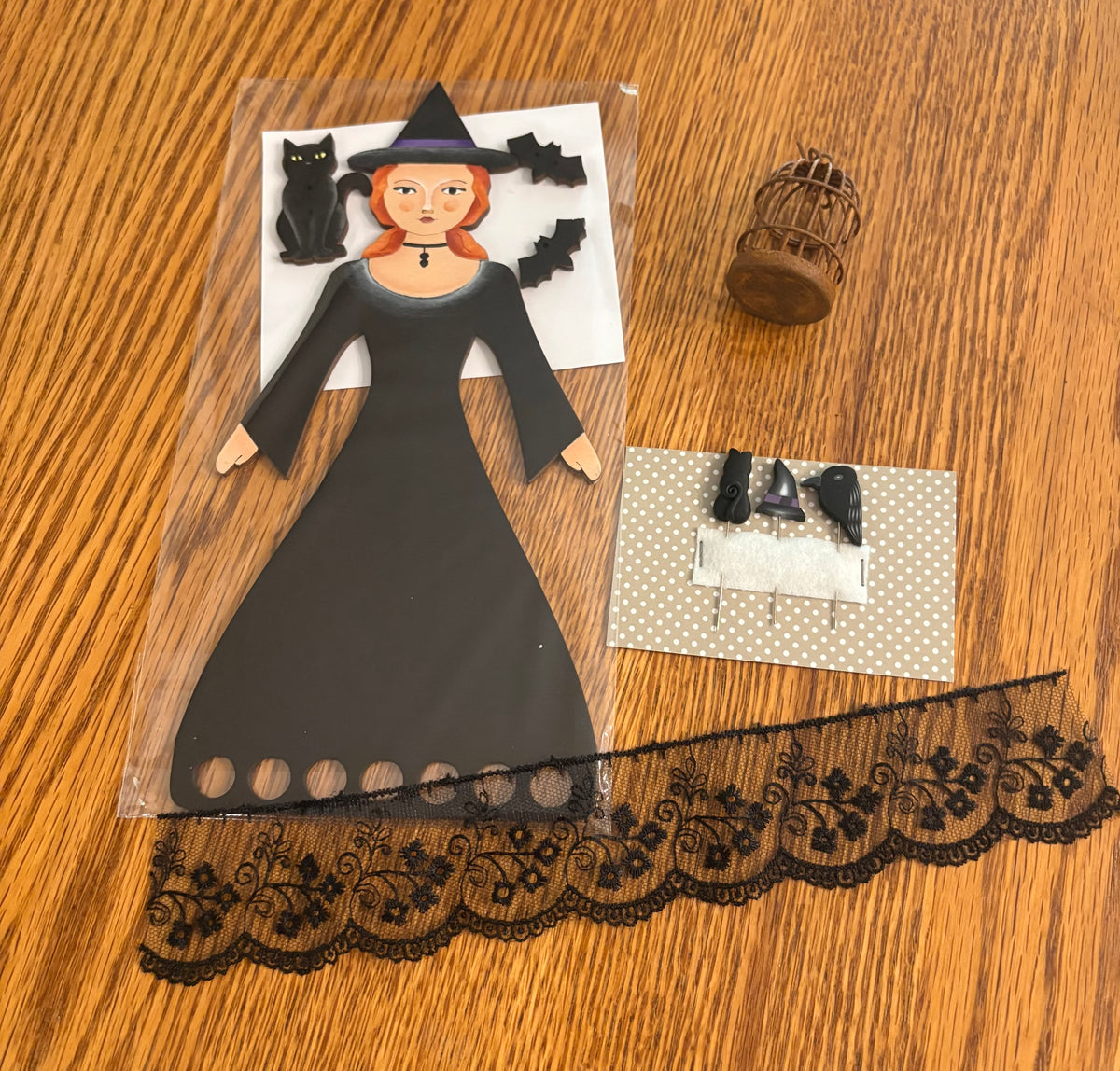 A Halloween-themed paper craft kit featuring a black witch dress with a printed face, black bats, a black cat, a small birdcage, and a black lace trim, laid out on a wooden table.