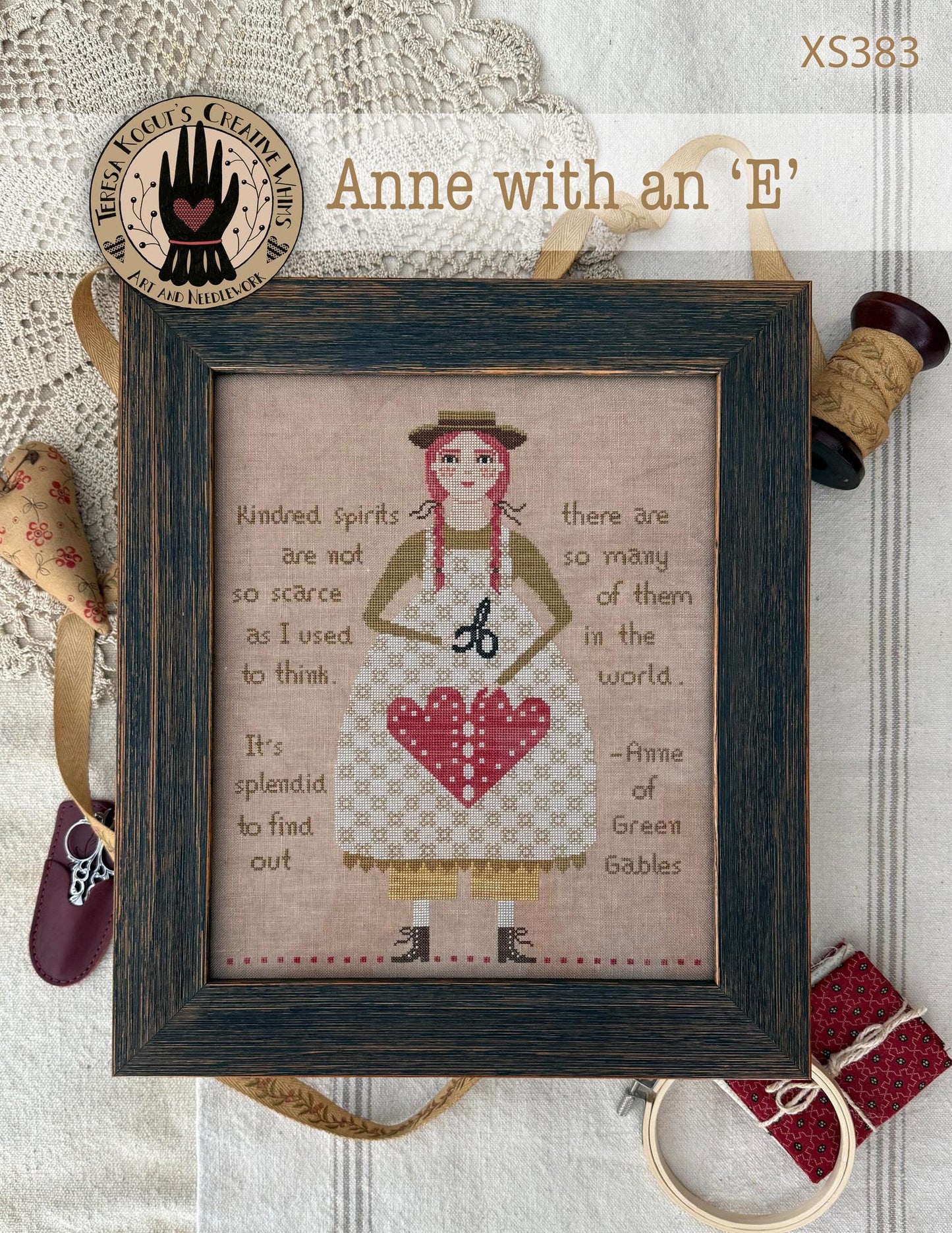 Framed cross-stitch pattern of Anne with an 'E' on a textured surface with sewing-themed items.