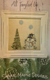 A framed needlepoint or cross-stitch design featuring a snowman and a Christmas tree on a beige fabric background, with blue snowflakes. The piece is displayed in a cream-colored frame against a rustic wood wall, with the text 'All Tangled Up' above and 'Sugar Maple Designs' at the bottom.