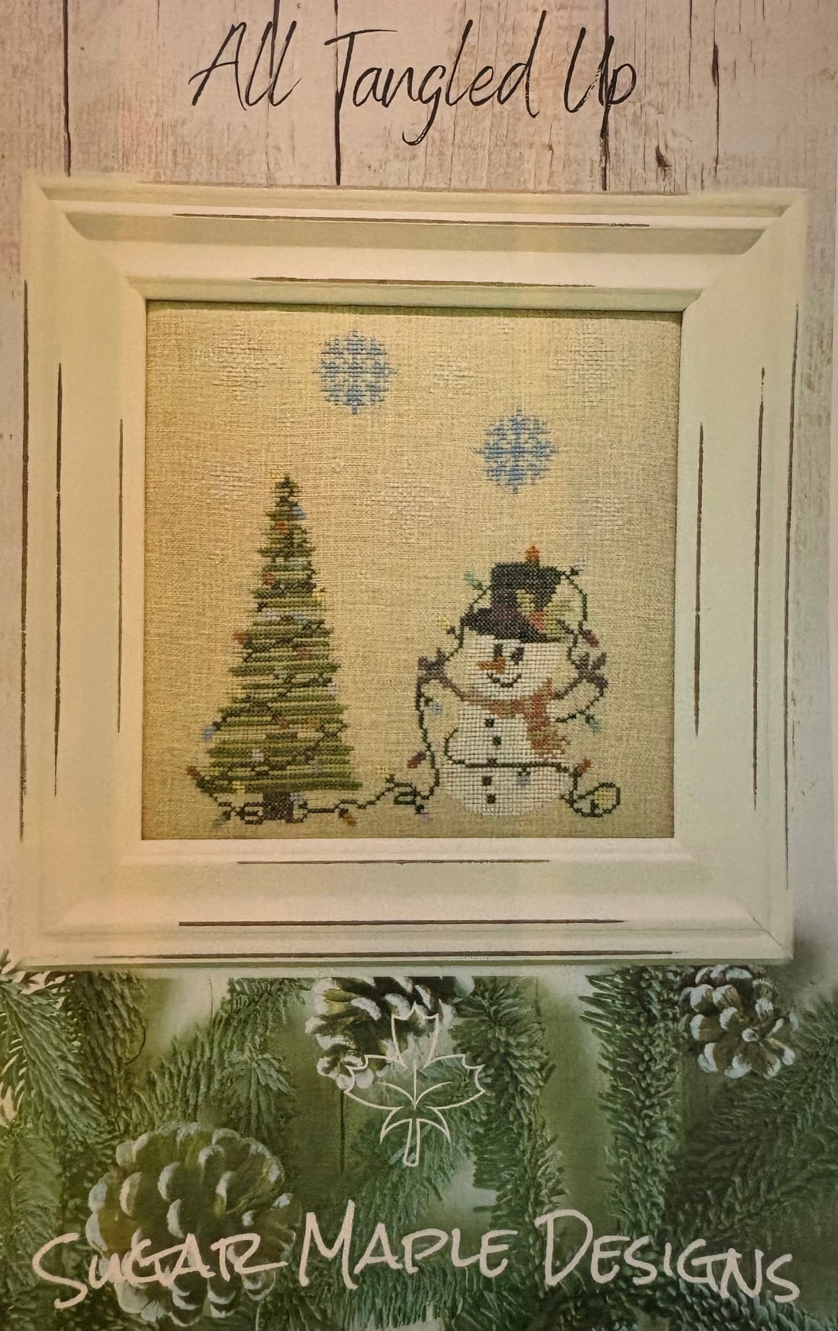 A framed needlepoint or cross-stitch design featuring a snowman and a Christmas tree on a beige fabric background, with blue snowflakes. The piece is displayed in a cream-colored frame against a rustic wood wall, with the text 'All Tangled Up' above and 'Sugar Maple Designs' at the bottom.