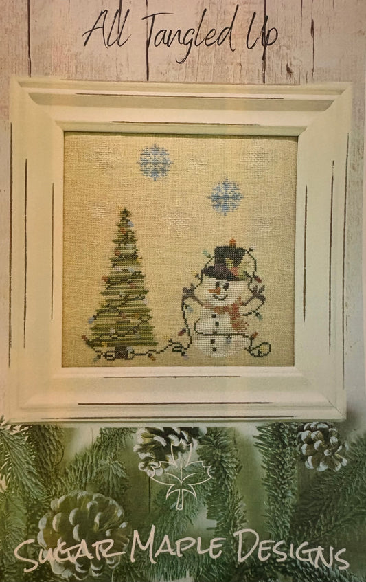 A framed needlepoint or cross-stitch design featuring a snowman and a Christmas tree on a beige fabric background, with blue snowflakes. The piece is displayed in a cream-colored frame against a rustic wood wall, with the text 'All Tangled Up' above and 'Sugar Maple Designs' at the bottom.