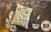 A square cross-stitch pillow with a blue and yellow floral design and the words 'Bee Happy' stitched in the center, displayed on a rustic wooden background with white daisies. The image includes 'Bee Happy' script at the top and a 'Summer House Stitches Works' logo at the bottom right.