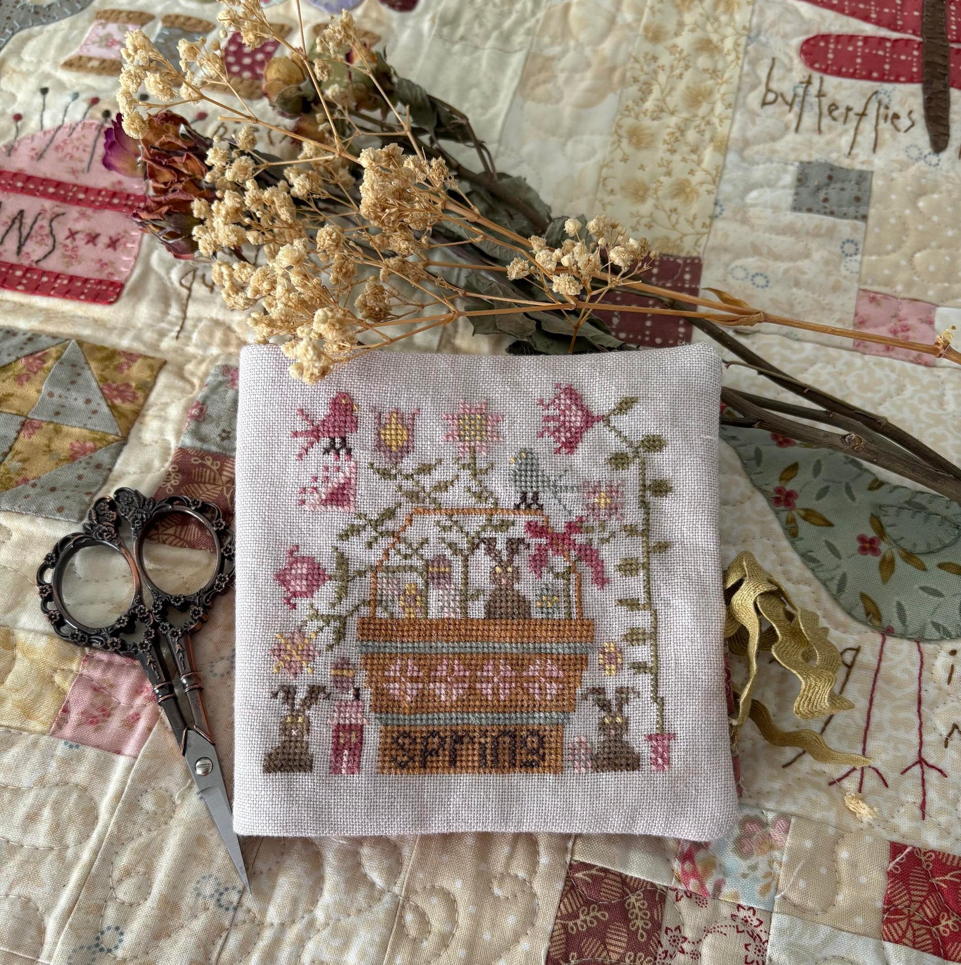 Cross-stitched piece with 'Spring' text on a patchwork background with dried flowers and scissors.