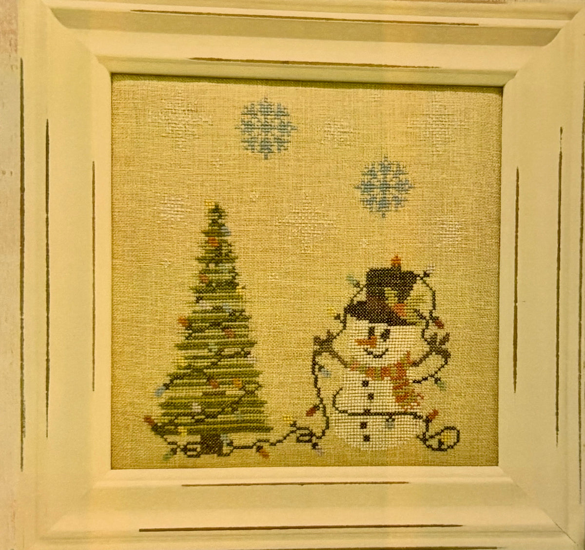 A framed needlepoint canvas featuring a Christmas tree and a snowman, set in a distressed cream-colored wooden frame.