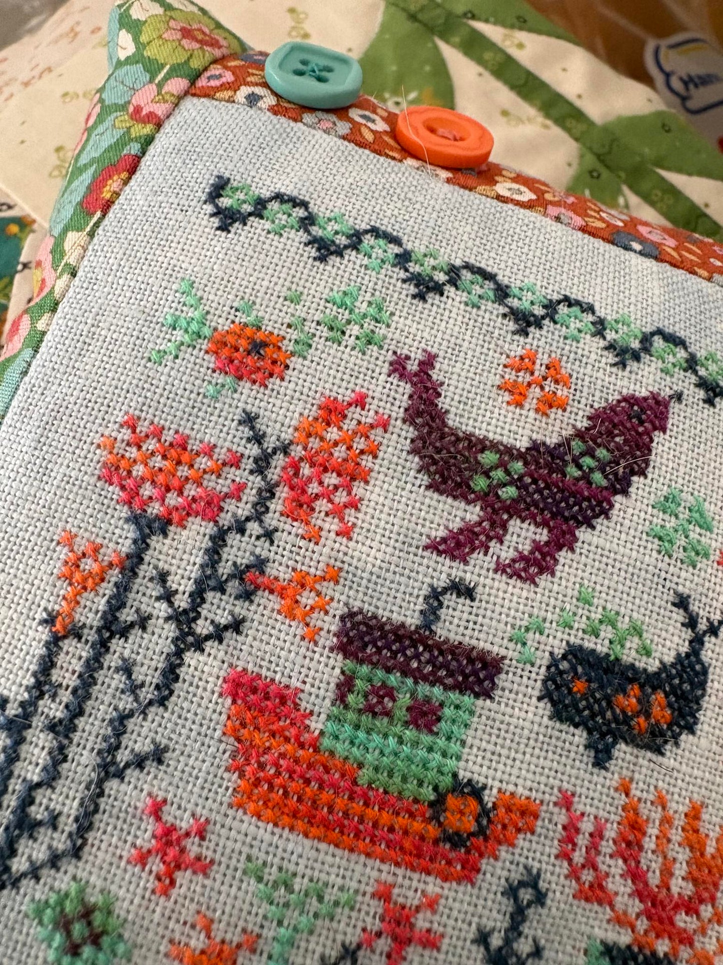 Cross-stitched piece with a bird, flowers, and a small house on fabric.