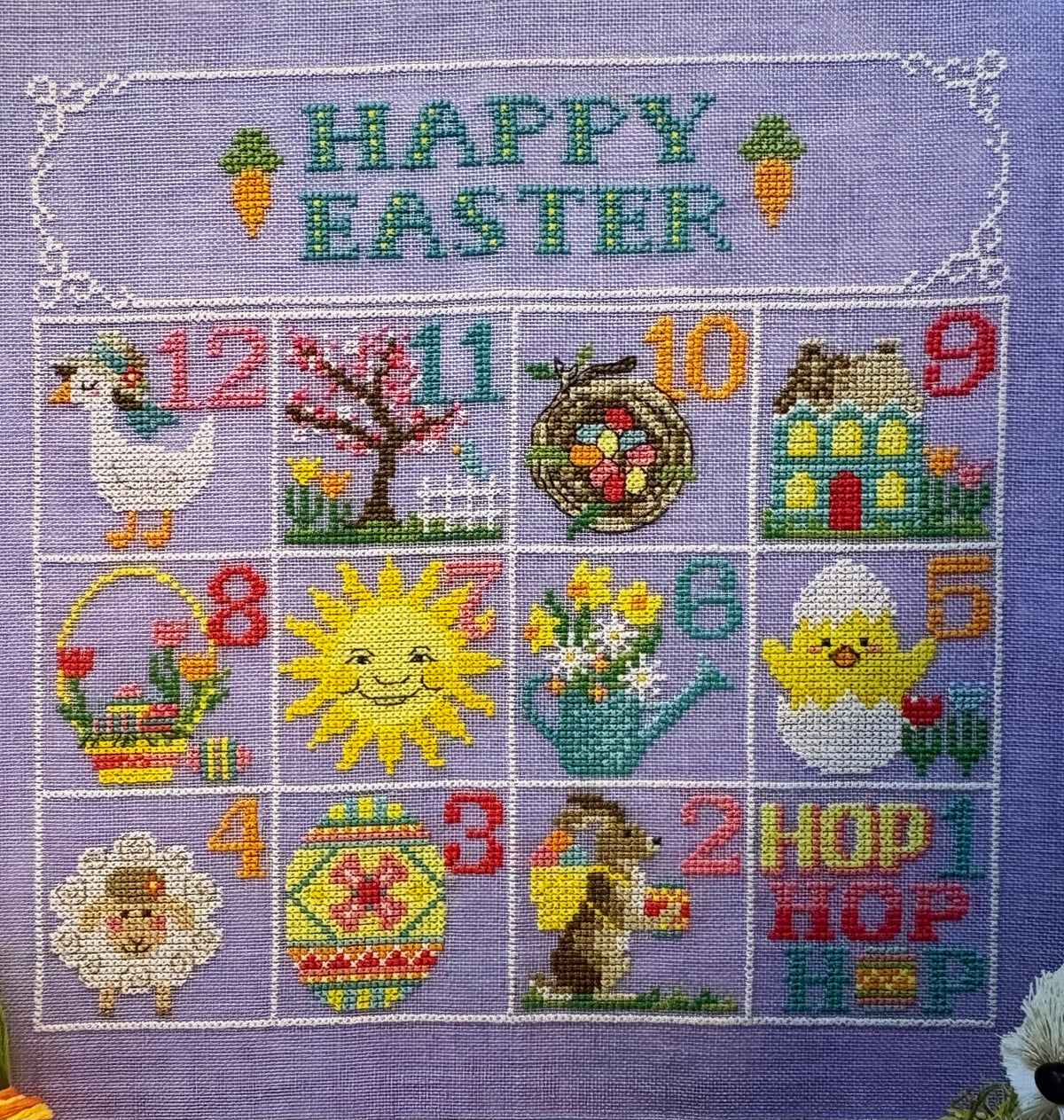 A square lavender cross-stitch sampler featuring an Easter-themed numbered grid from 1 to 12, with 'HAPPY EASTER' stitched at the top. The design includes colorful motifs such as a duck, tree, basket, sun, flowers, house, sheep, egg, bunny, and chick, all on a purple fabric background.