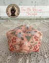 Decorative biscornu with floral patterns on a textured surface, featuring a brand logo in the corner.
