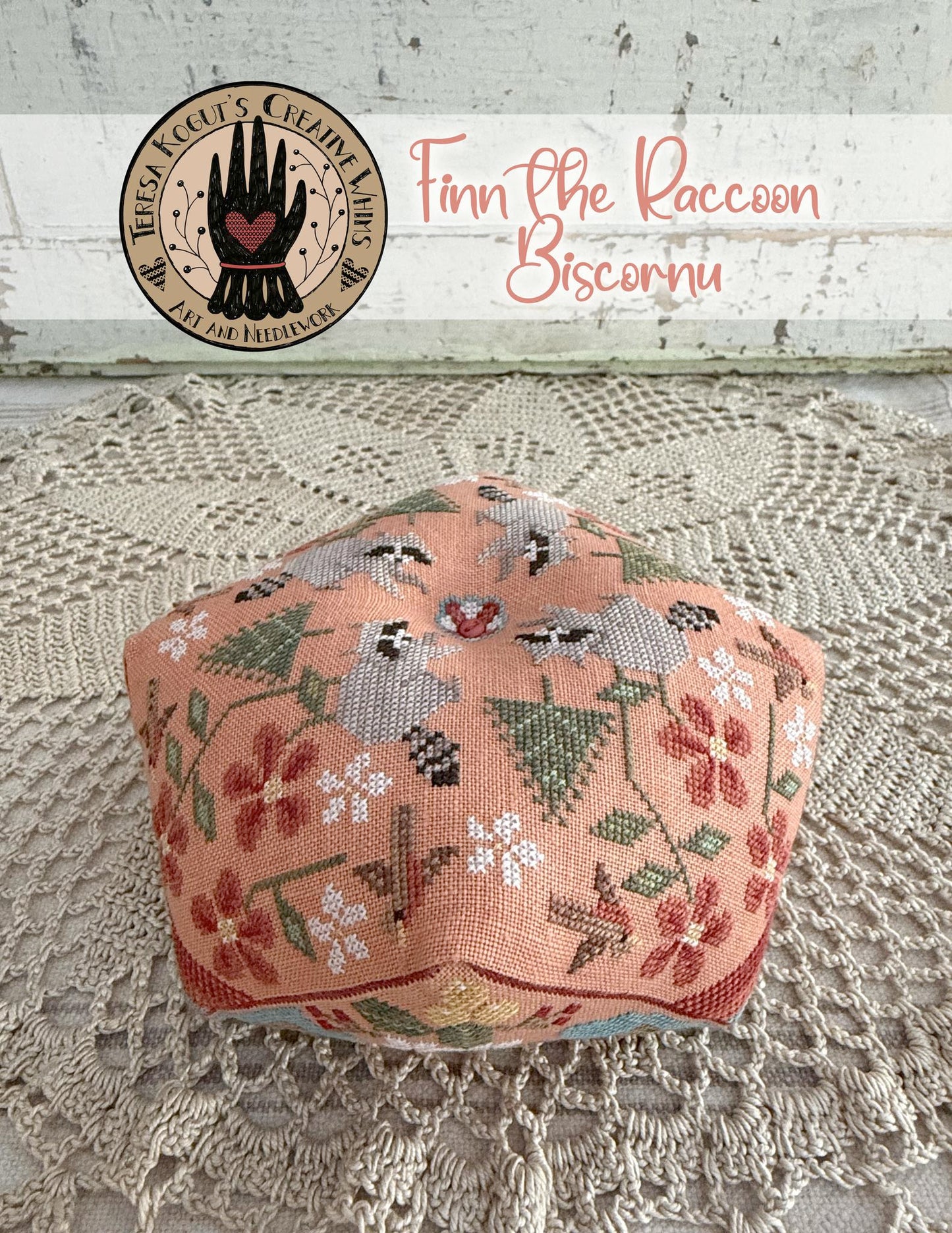 Decorative biscornu with floral patterns on a textured surface, featuring a brand logo in the corner.