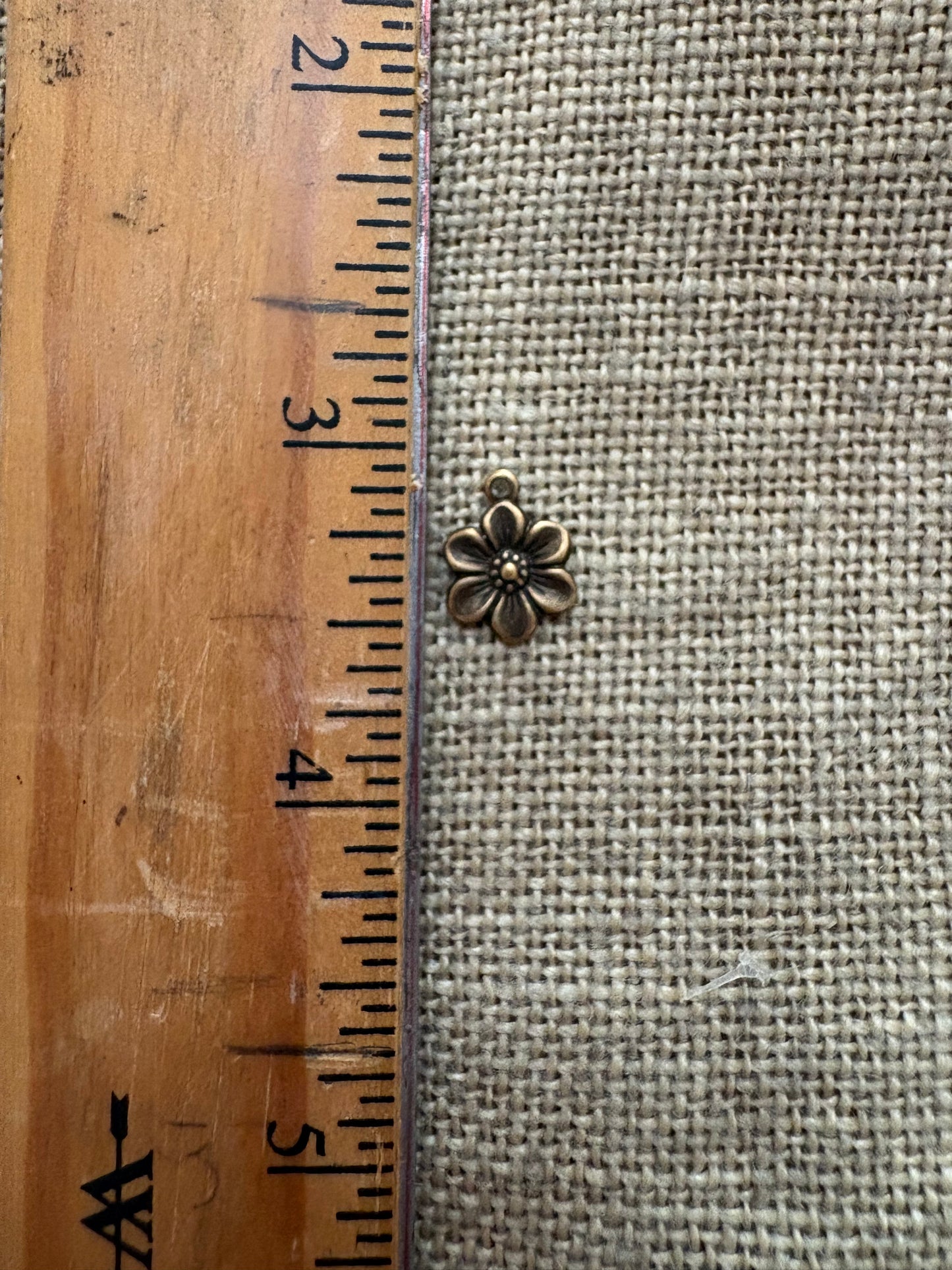 Small metal flower charm next to a wooden ruler on a textured fabric background