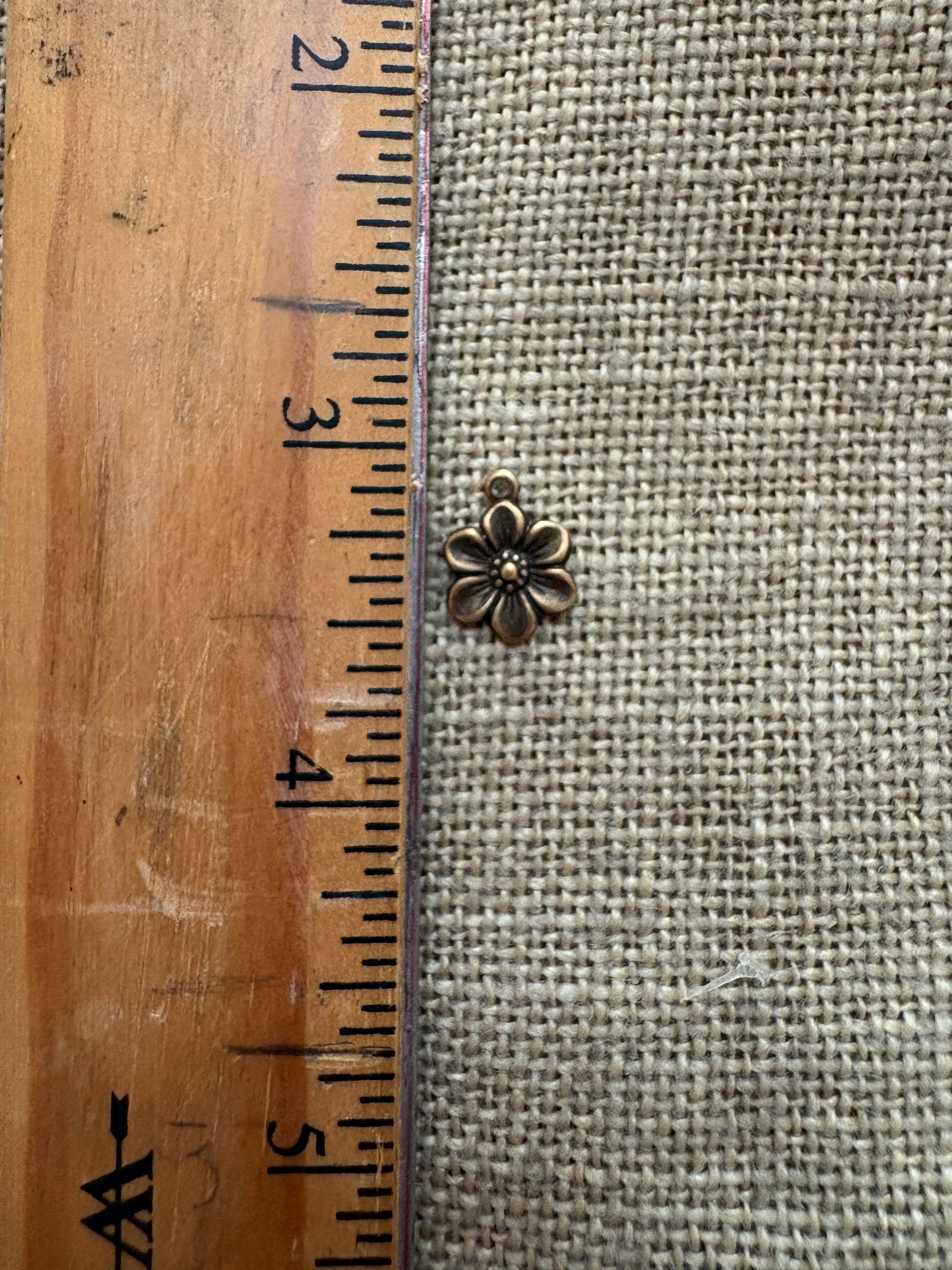 Small metal flower charm next to a wooden ruler on a textured fabric background
