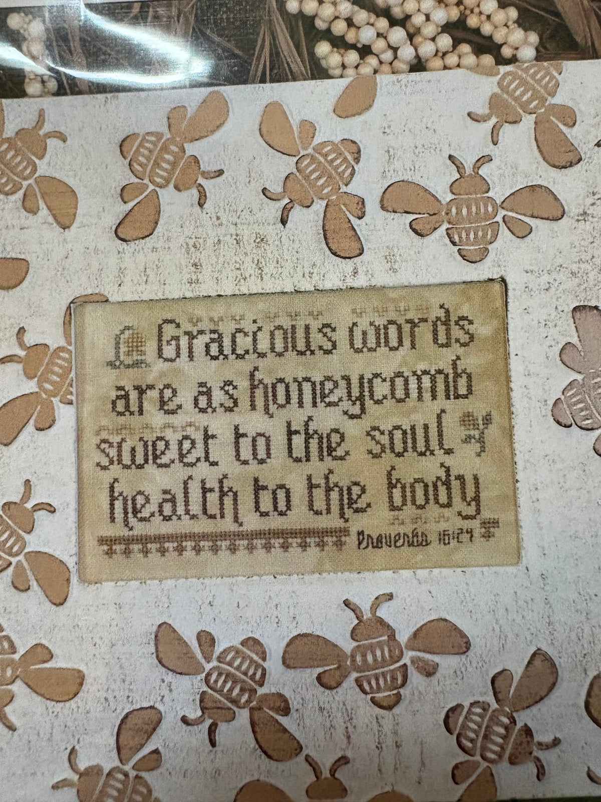 A close-up of a decorative craft or gift item featuring a central beige patch with the phrase, 'Gracious words are as honeycomb sweet to the soul, health to the body' and 'Proverbs 16:24' at the bottom. The surrounding area is decorated with laser-cut wooden bees on a light beige textured background.