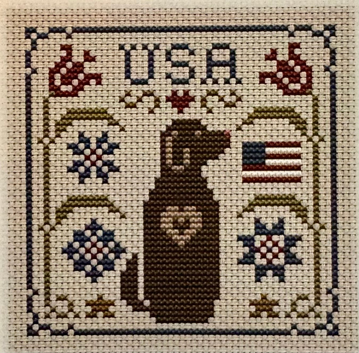 A square cross-stitch sampler featuring a brown dog silhouette with a heart on its chest, the letters USA at the top, an American flag motif, and decorative floral and geometric patterns on a cream fabric background.