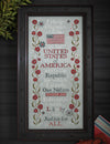 Framed cross-stitched piece with the Pledge of Allegiance and American flag on a wooden background.