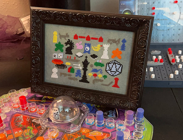 Framed cross-stitch art of colorful game pieces on a table with gaming supplies.