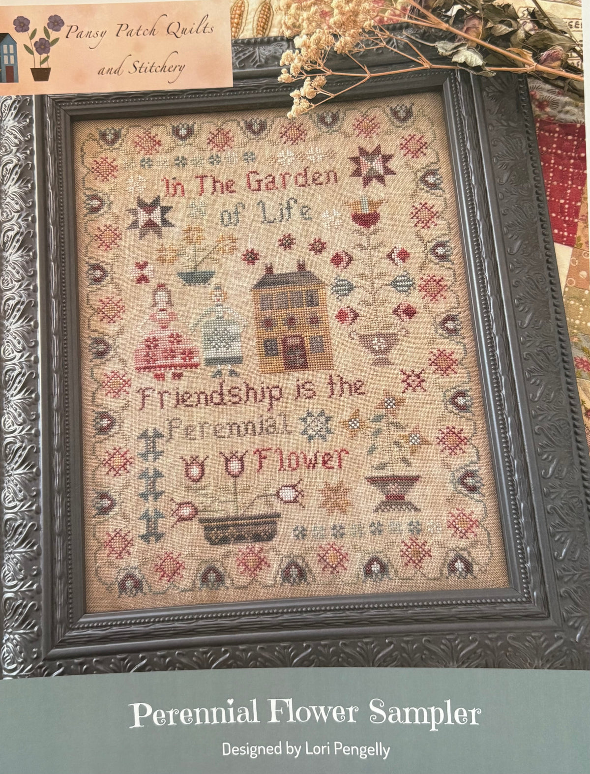 A framed cross-stitch sampler titled 'Perennial Flower Sampler' with the stitched design reading 'In the Garden of Life' and 'Friendship is the Perennial Flower.' The sampler features a beige linen background with multicolored floral and house motifs, enclosed in a dark ornate frame.
