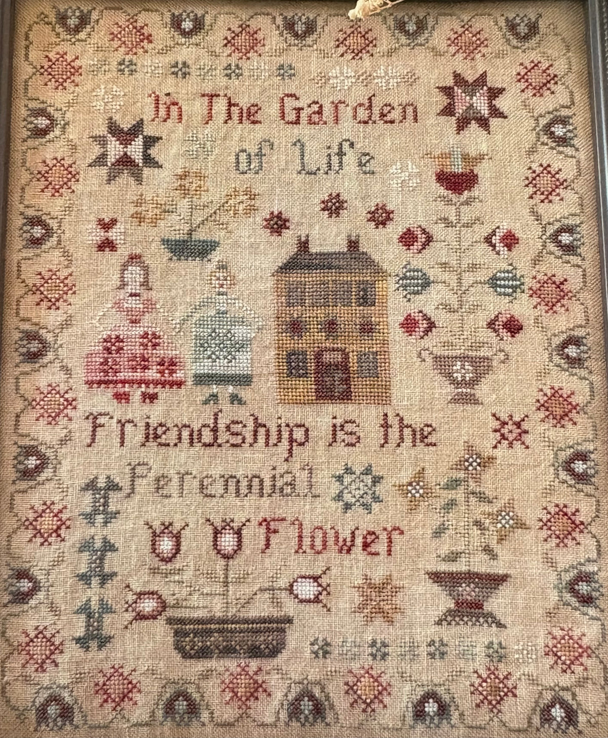A framed needlepoint sampler with a rustic folk-art design. The beige linen background features stitched text reading 'In The Garden of Life' and 'Friendship is the Ferennial Flower,' surrounded by motifs of a house, flowers, stars, and geometric borders in muted red, brown, gray, and green tones.