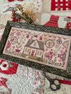Framed cross-stitched piece with a house design on a quilt background