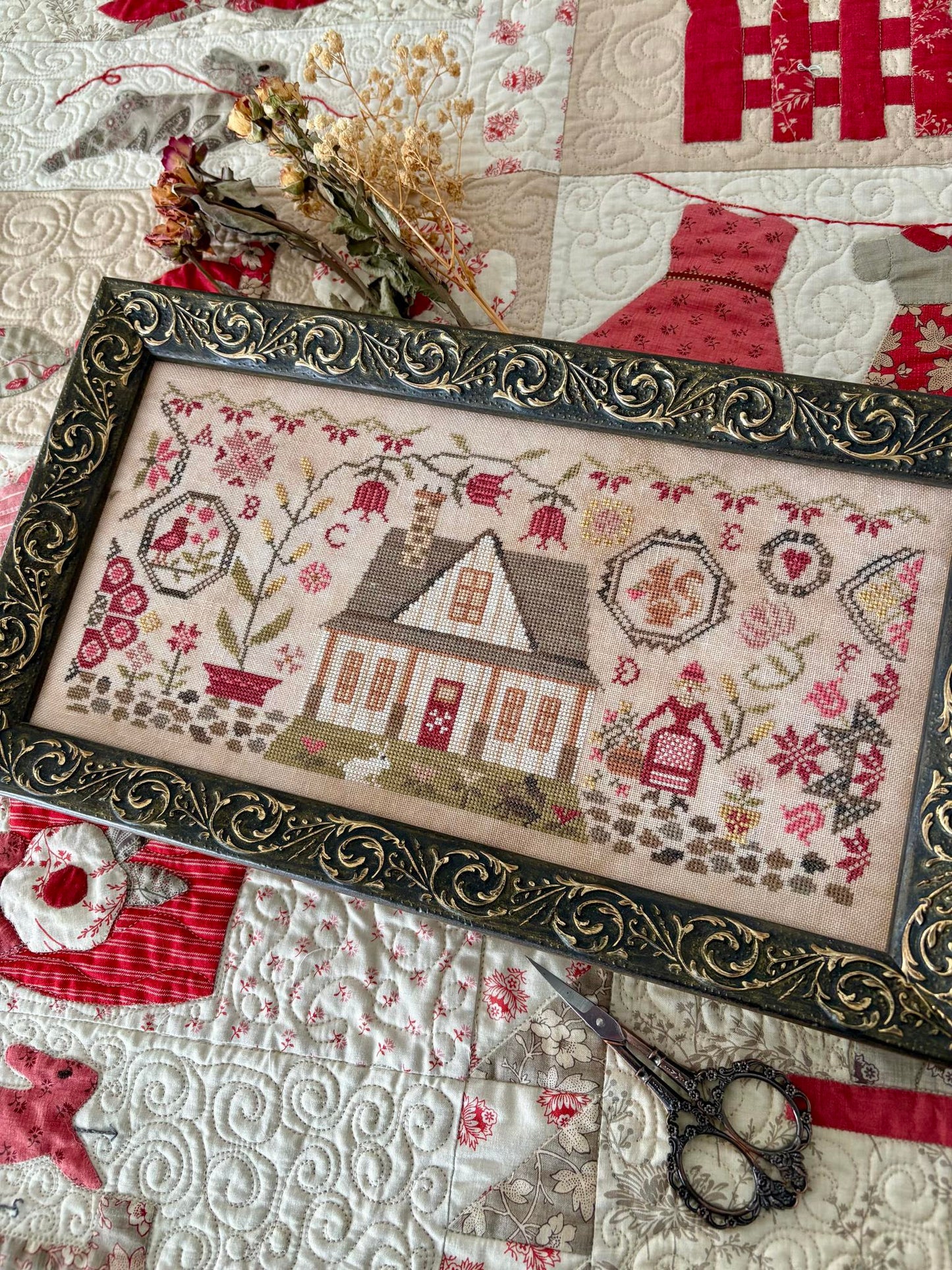 Framed cross-stitched piece with a house design on a quilt background