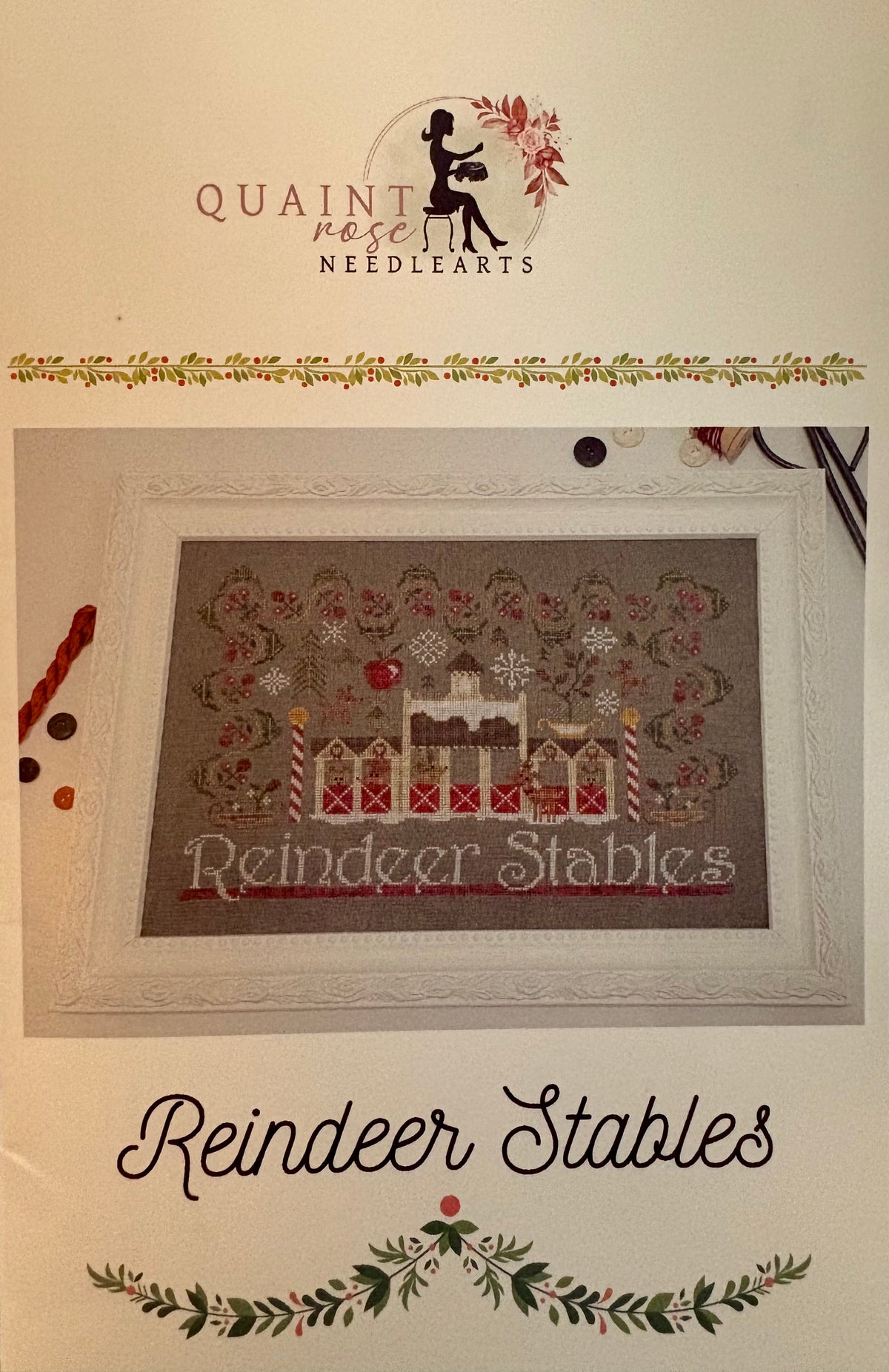 A framed cross-stitch design reading 'Reindeer Stables' shown on a cream background with the Quaint Rose Needlearts logo at the top and the title 'Reindeer Stables' at the bottom.