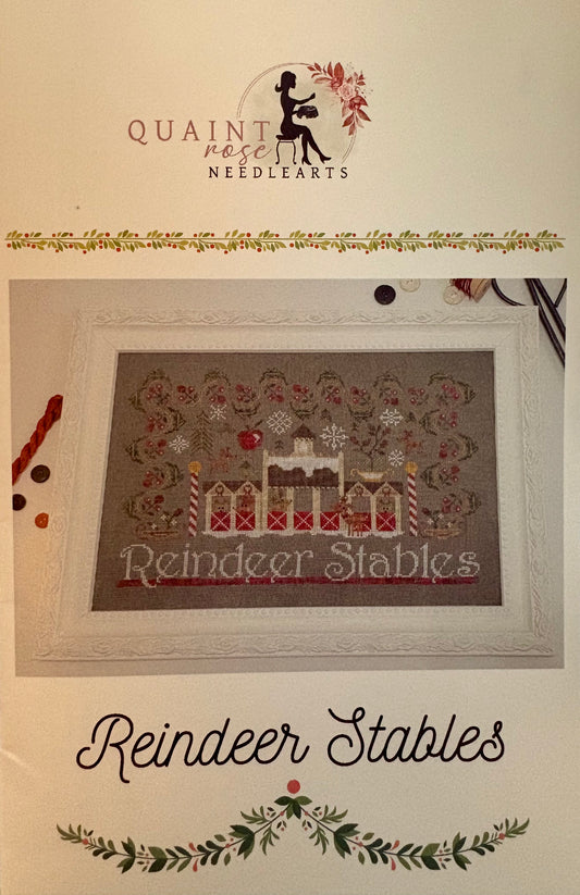 A framed cross-stitch design reading 'Reindeer Stables' shown on a cream background with the Quaint Rose Needlearts logo at the top and the title 'Reindeer Stables' at the bottom.