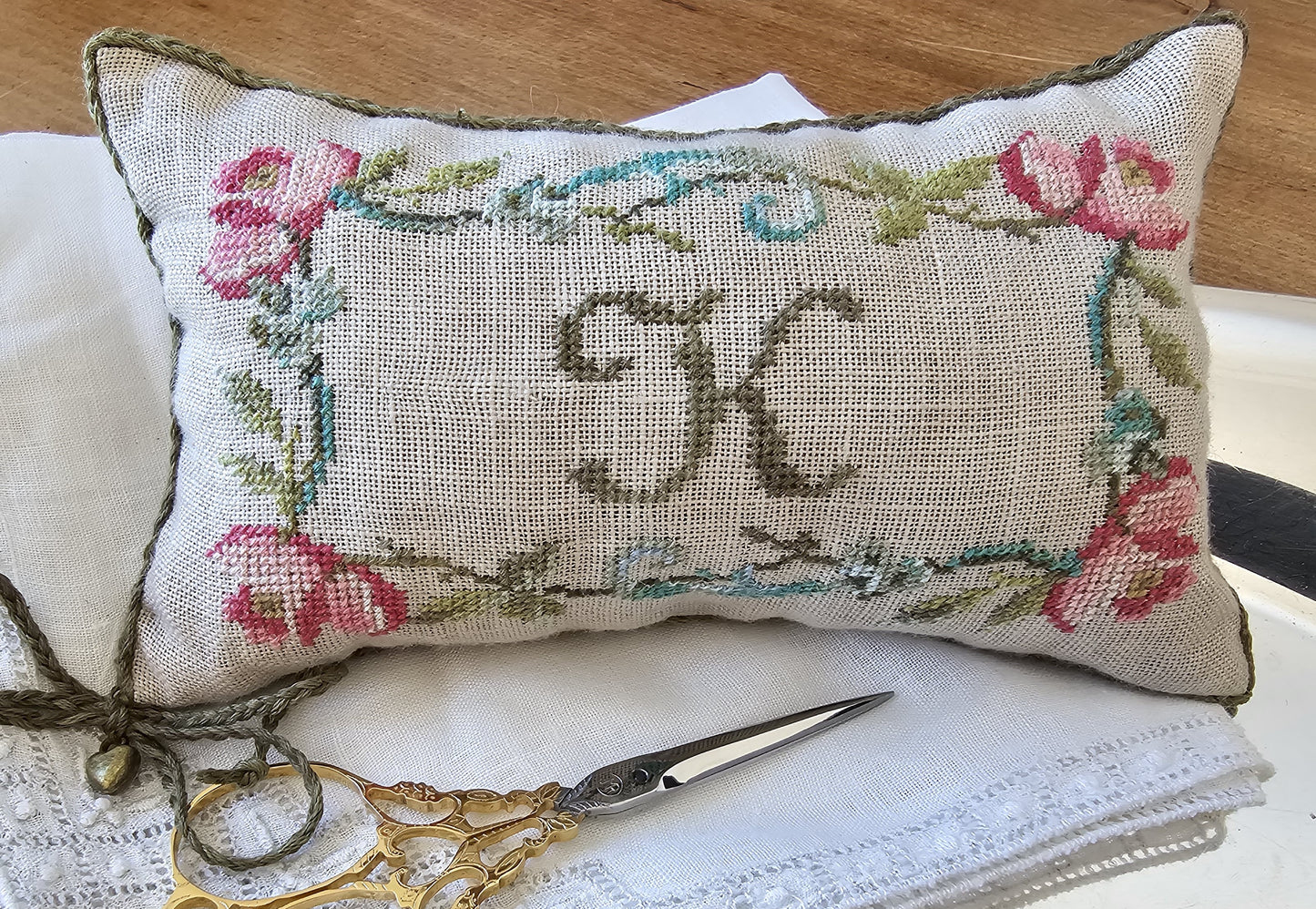 Decorative pillow with floral patterns and initials, accompanied by a pair of scissors on a wooden surface.