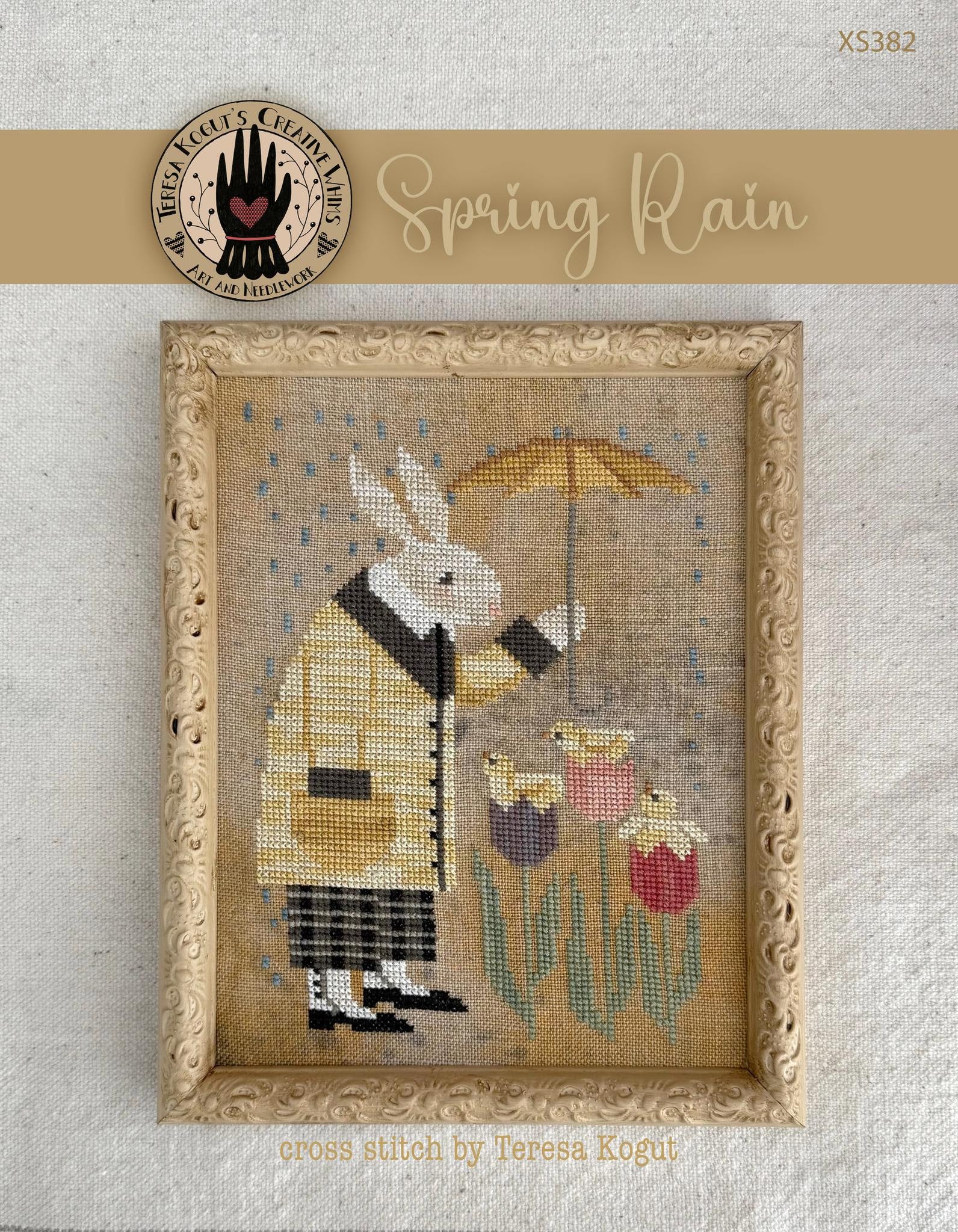 Cross-stitched design of a rabbit holding an umbrella with a chick, framed in gold on a textured beige background.
