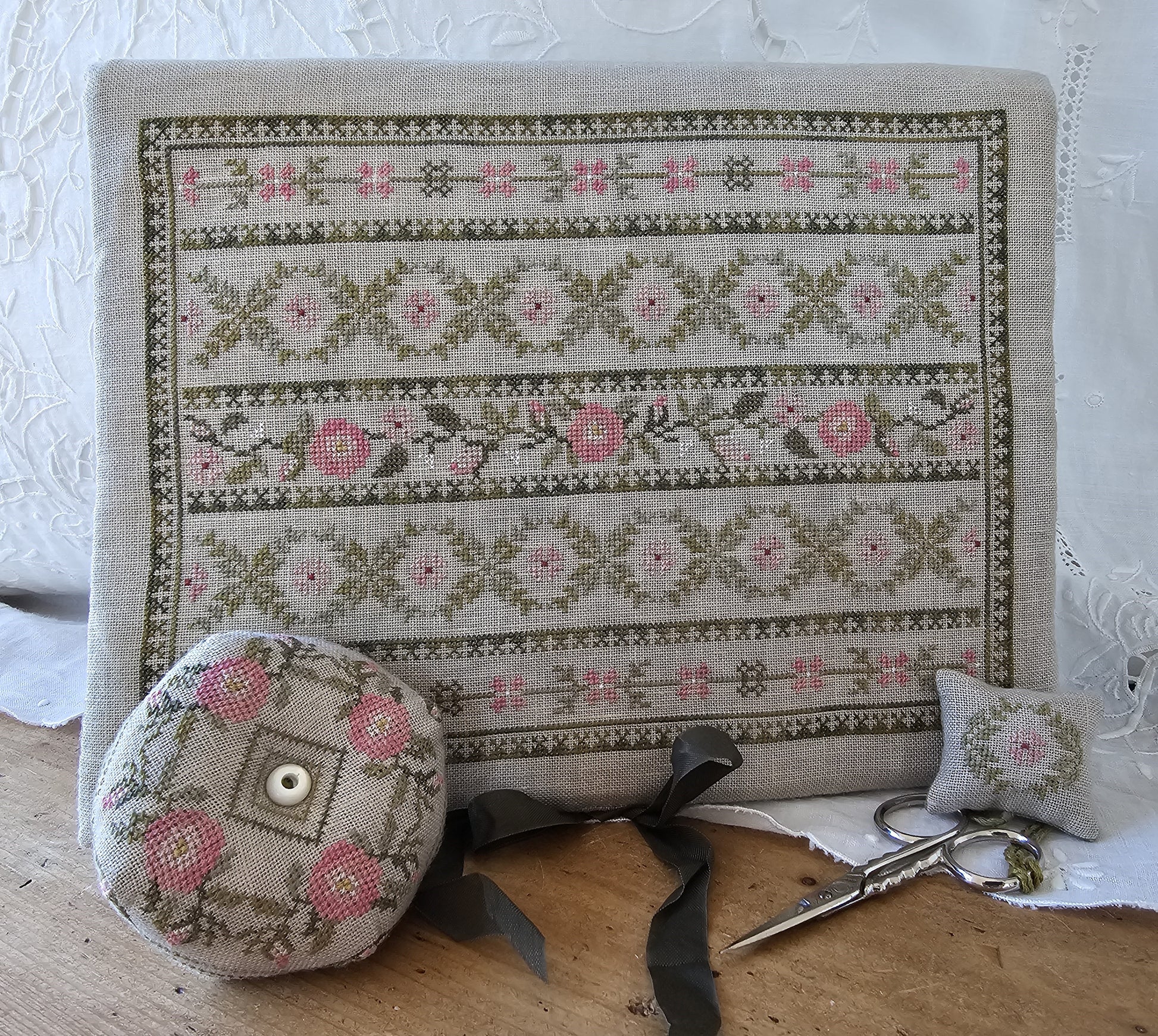 Cross-stitched fabric with floral patterns, a pin cushion, scissors, and a thimble on a wooden surface.