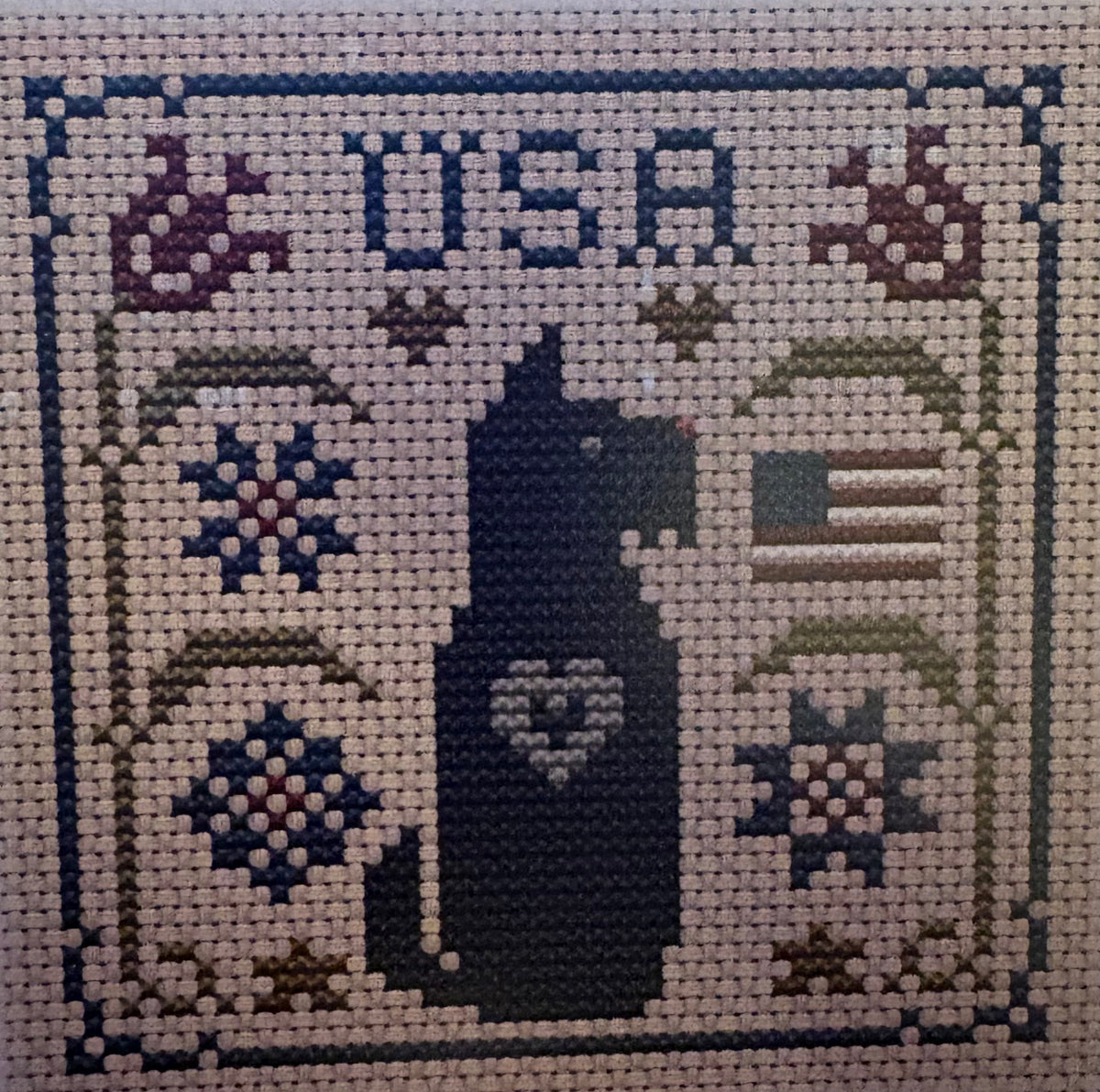 A square cross-stitch sampler featuring a dark blue silhouette of a dog with a heart on its chest, surrounded by floral motifs, the letters 'USA' at the top, and a small American flag on the right.