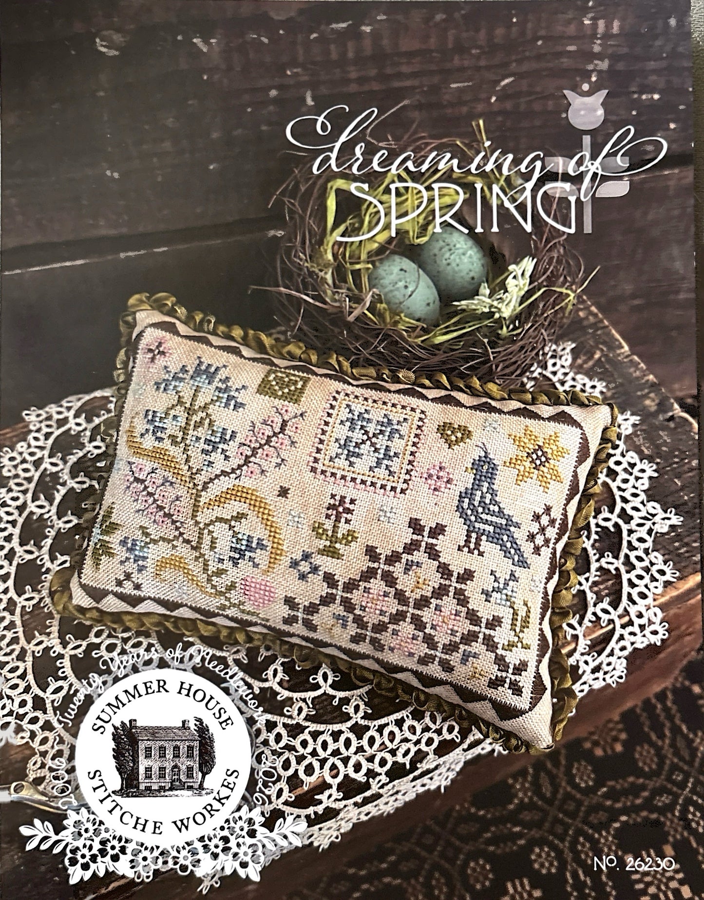 Cross-stitched pillow with decorative patterns, eggs in a nest, and 'Dreaming of Spring' text on a wooden surface.