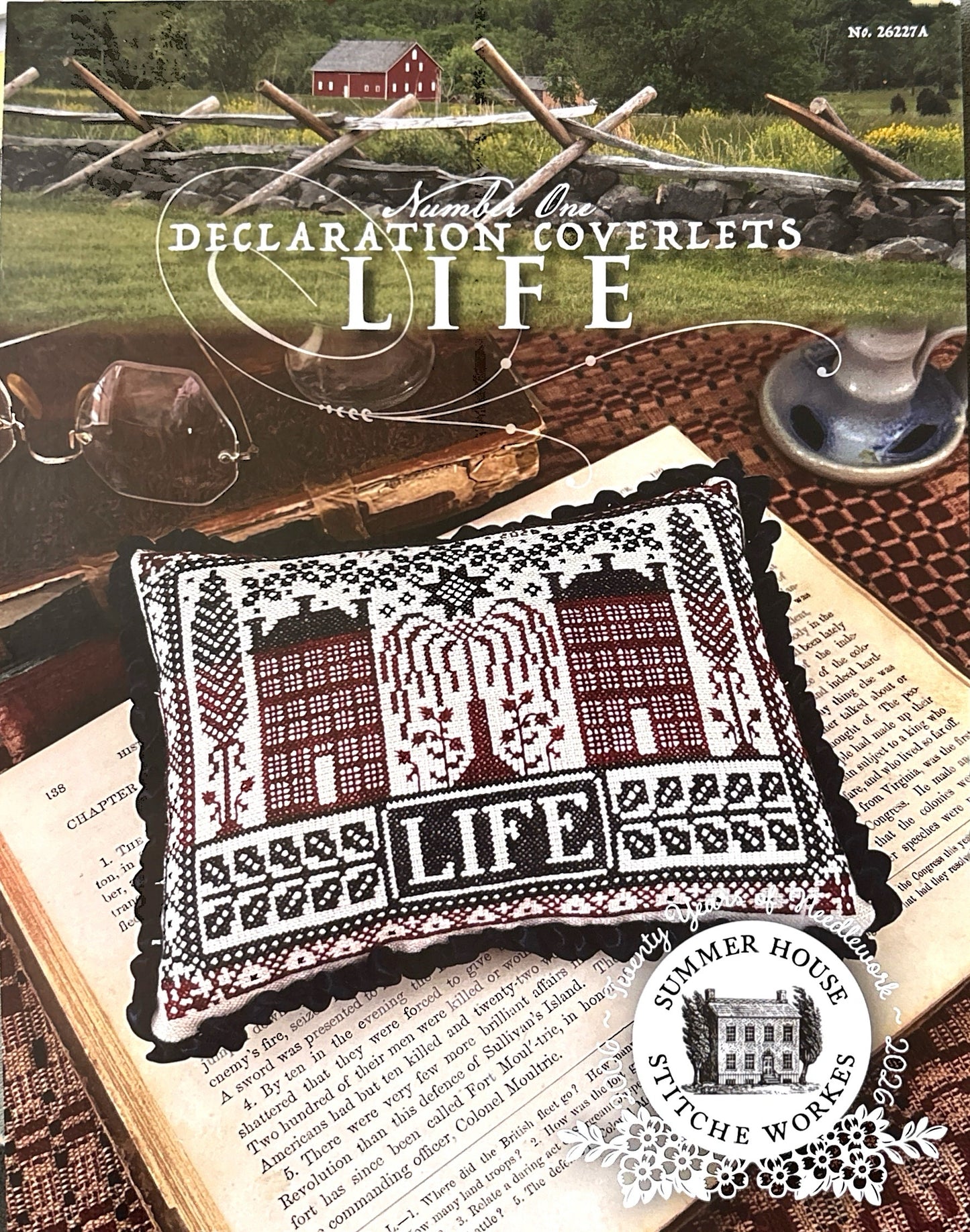 Cross-stitched pillow with 'LIFE' design on an open book, surrounded by rustic elements.