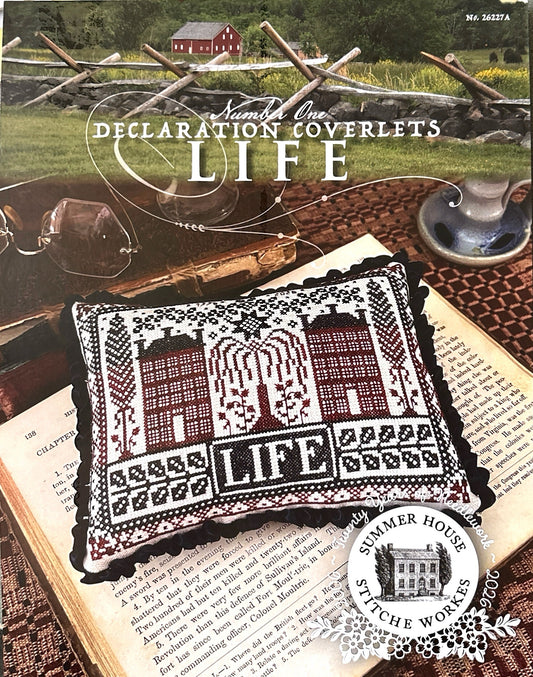 Cross-stitched pillow with 'LIFE' design on an open book, surrounded by rustic elements.
