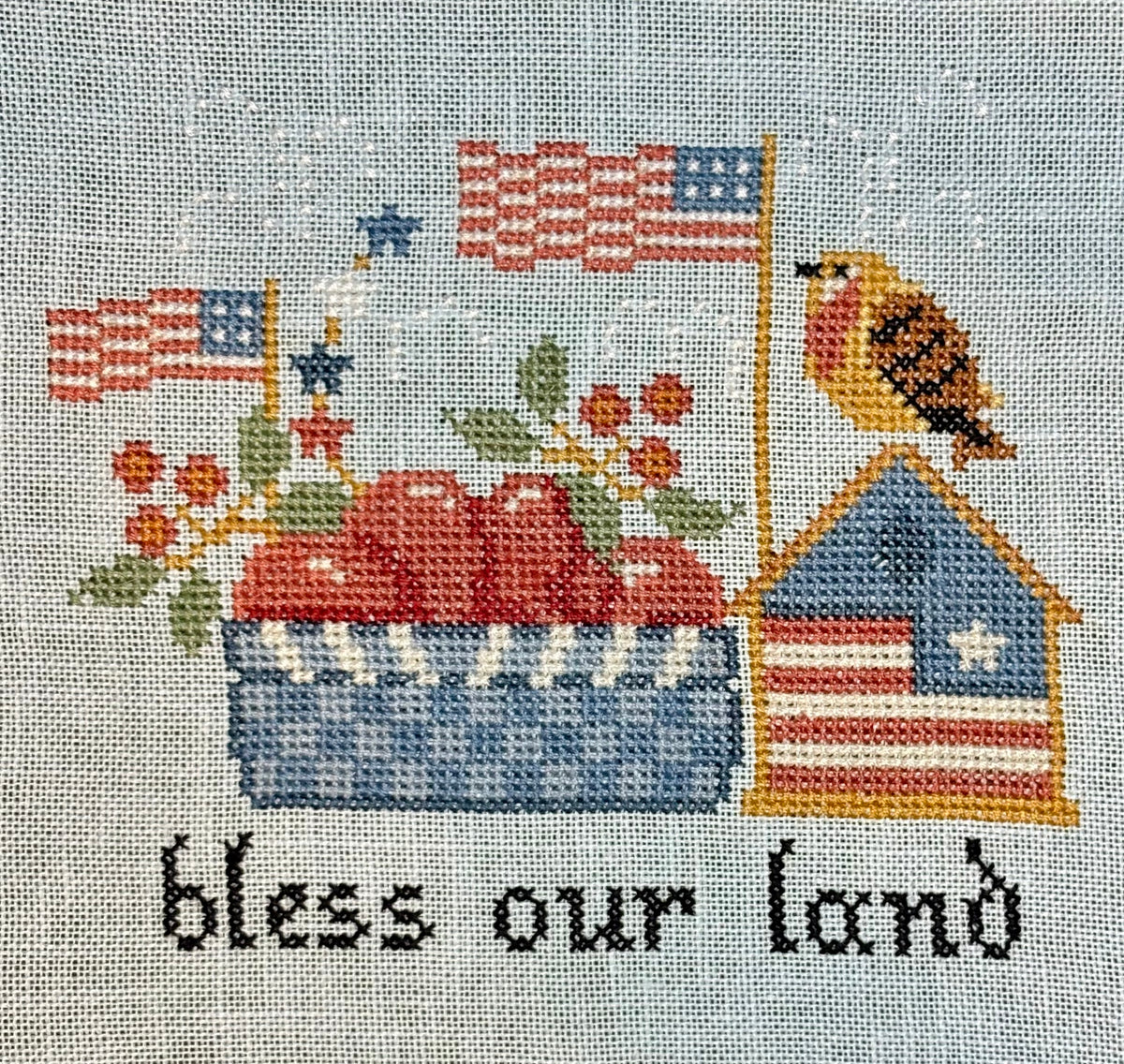 Embroidery design with American flags, a house, and text 'bless our land' on a fabric background.