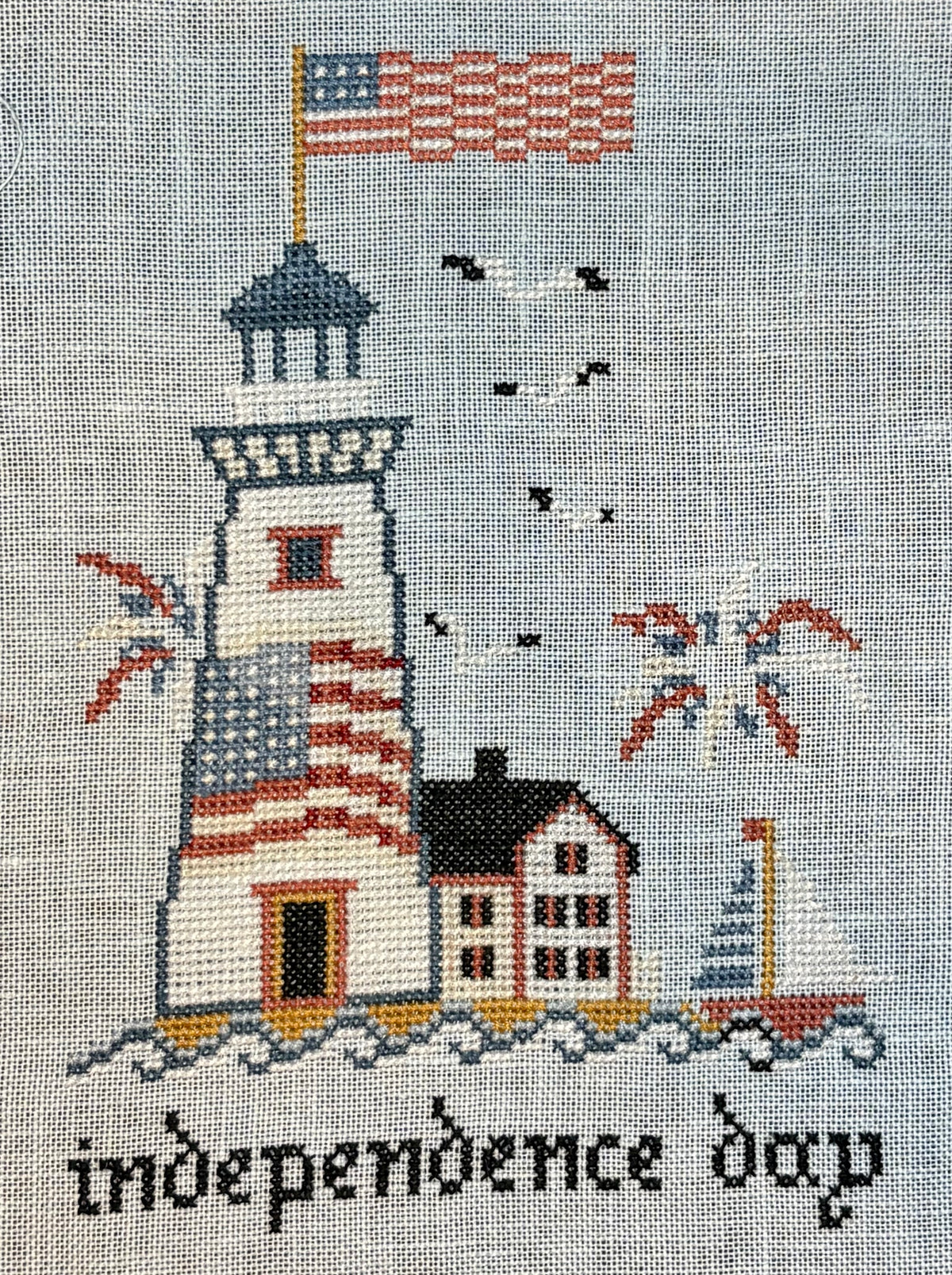 Cross-stitched design of a lighthouse with an American flag and 'independence day' text on a gray background.