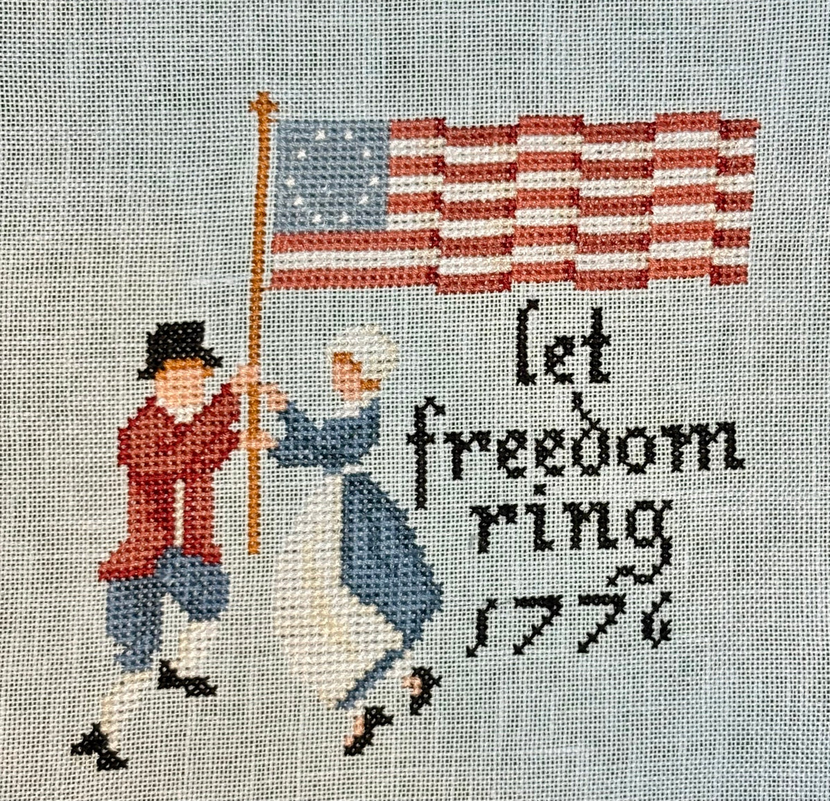 Cross-stitched design with two figures holding an American flag and the text 'let freedom ring 1776' on a gray background.
