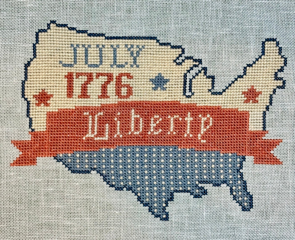 Cross-stitched map of the United States with 'Liberty' and '1776' text on a textured fabric background.