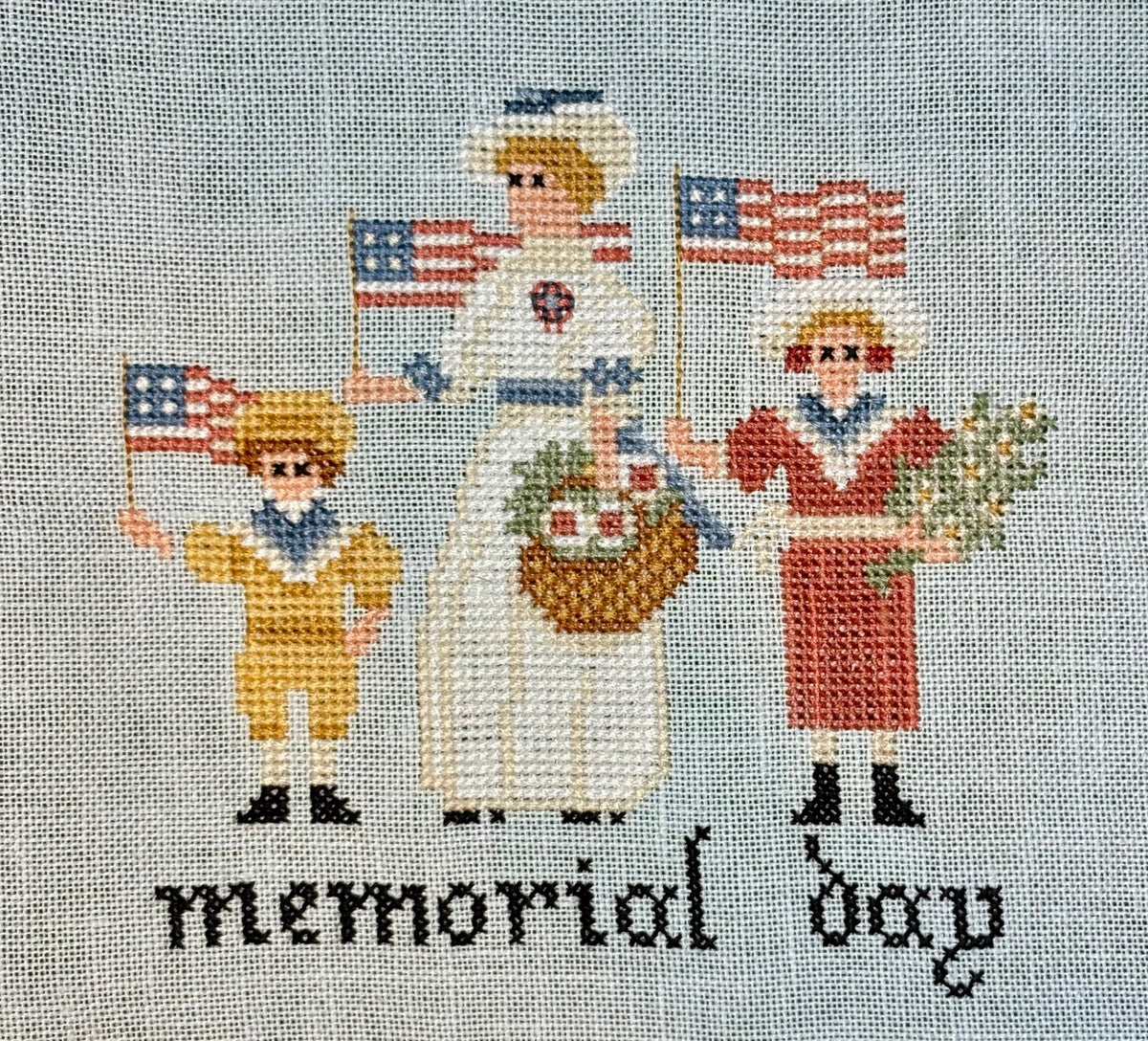 Cross-stitched design of a family holding American flags and wreaths with 'memorial day' text on a textured fabric background.