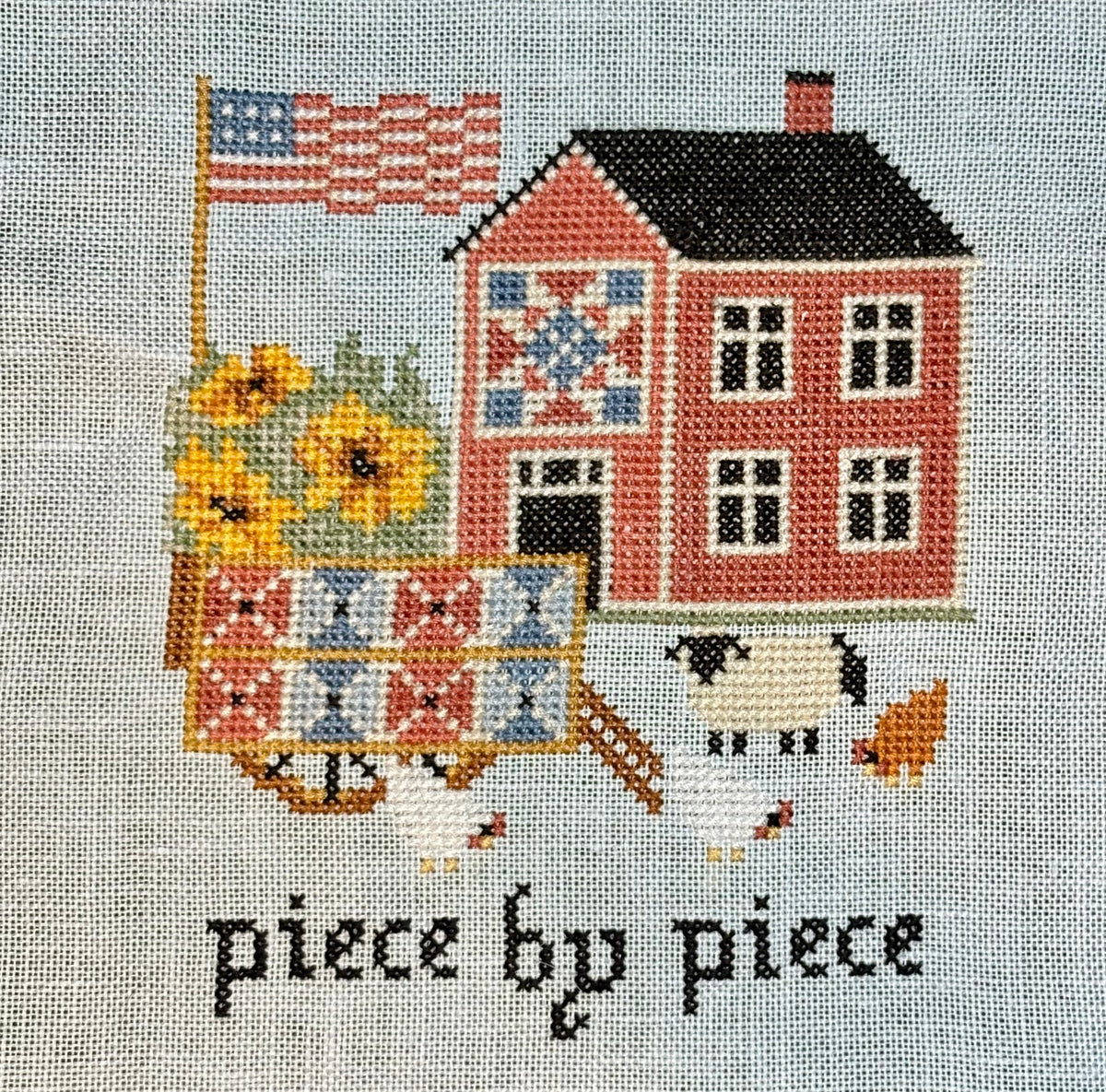 Cross-stitched design of a house with a quilt, sunflowers, and an American flag on fabric.