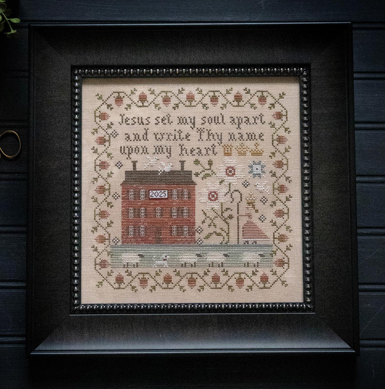 Framed cross-stitched piece with religious text and house illustration on a dark background